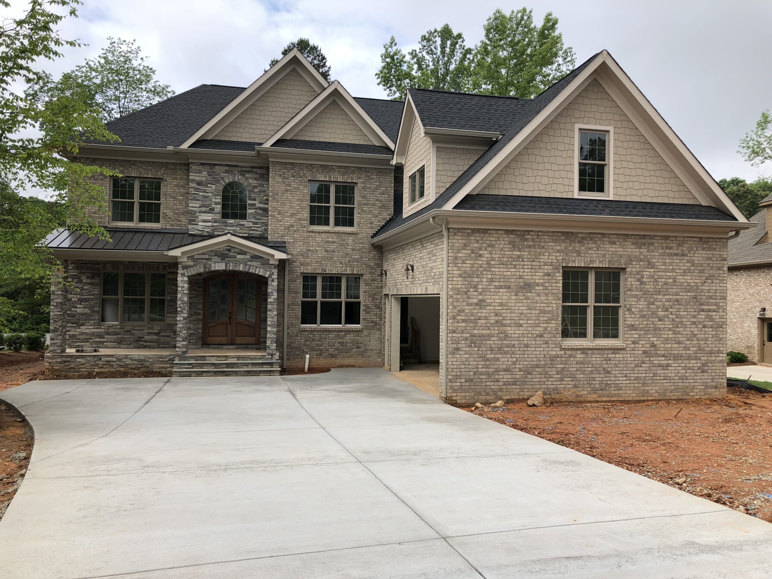 Photo May 25 2018, 9 31 57 AM Two-story house with a mix of brick and stone facade, double-front doors, and an attached garage on a freshly paved driveway. Surrounding trees in the background.