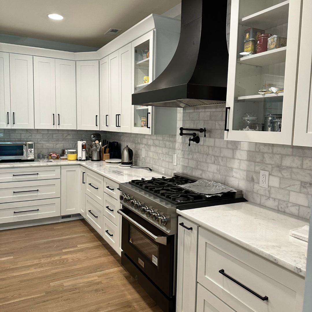 Modern kitchen with white cabinets, stainless steel appliances, a black range hood, marble backsplash, and various small kitchen items on the counters.
