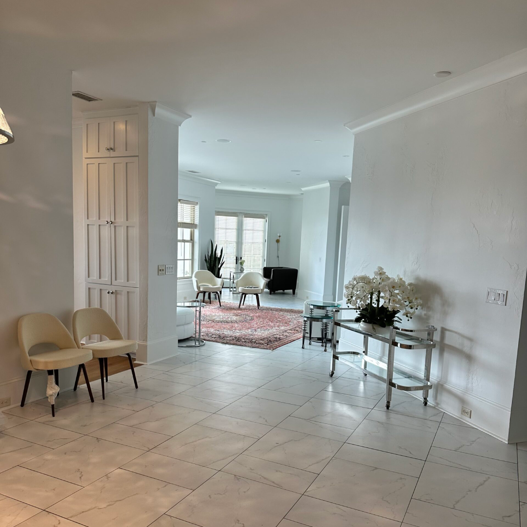 A bright, modern living space with white walls, tiled floor, neutral furnishings, a large window, and decorative plants and flowers.