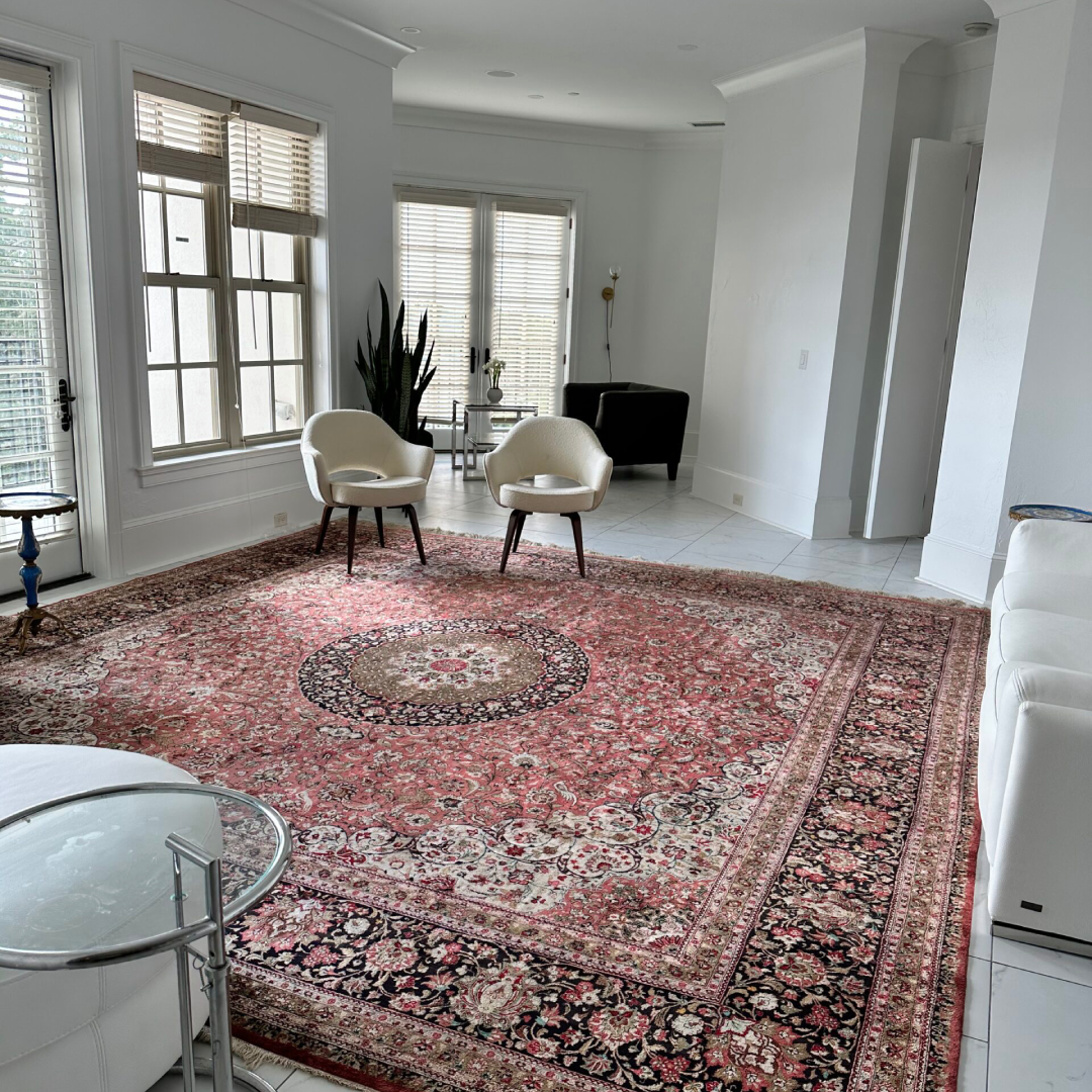 A spacious living room with a large patterned rug, two white chairs, a white sofa, a glass side table, a plant, and tall windows letting in natural light.