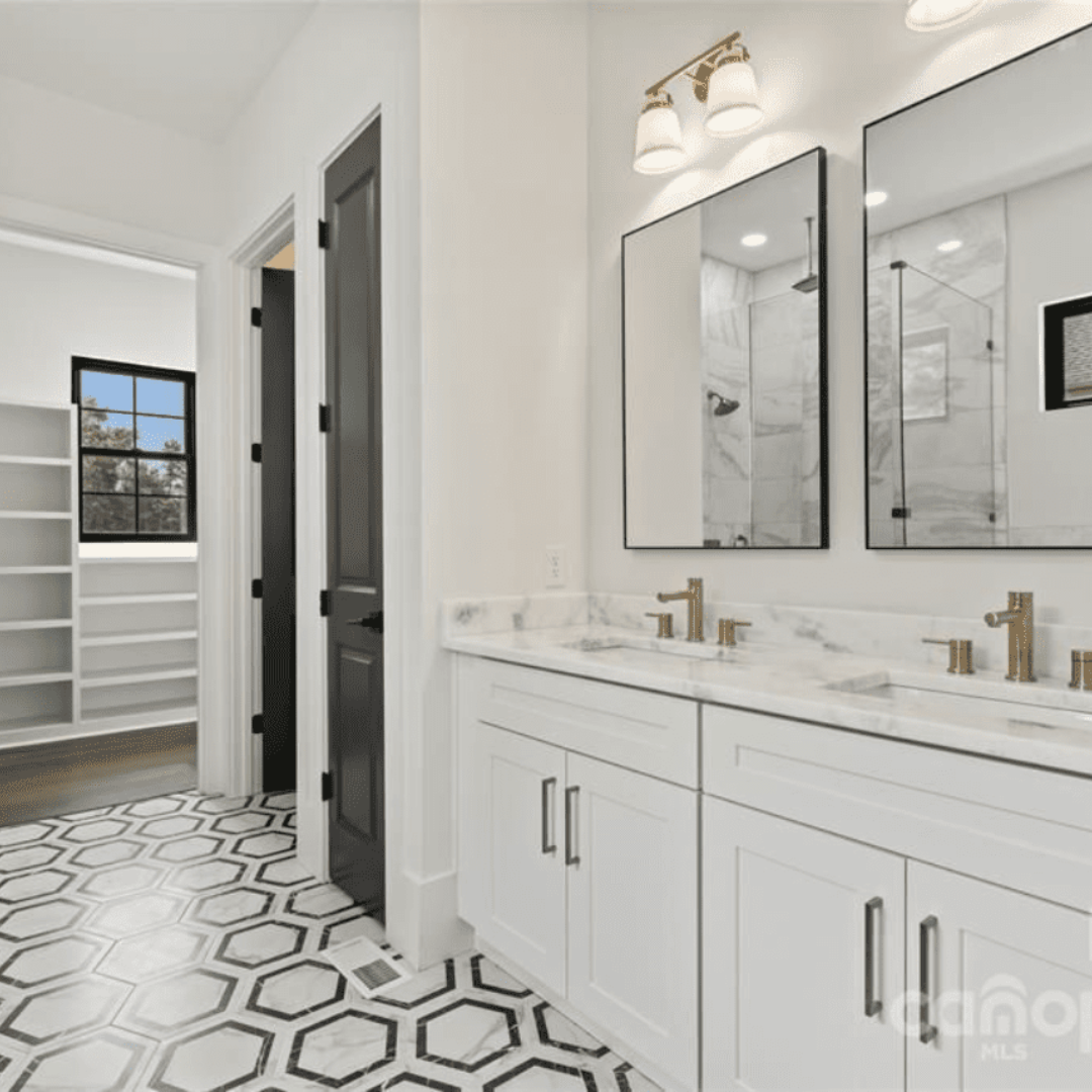 Modern bathroom with white double-sink vanity, marble countertop, two mirrors, black hexagonal tile floor, and a view into a walk-in closet with built-in shelves.