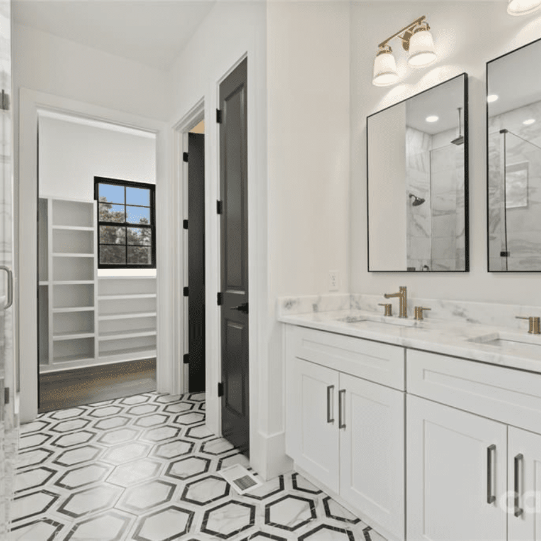 Modern bathroom with a double sink vanity, hexagonal tile floor, two mirrors, and brass fixtures; a walk-in closet with shelves is visible through an open doorway.